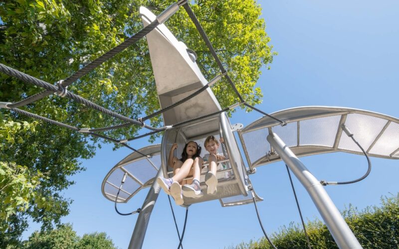 Zwei Kinder sitzen in einem an Seilen aufgehängten Spielplatz in Form eines Metallflugzeugs, mit Bäumen und einem klaren blauen Himmel im Hintergrund.