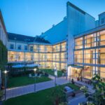 Ein modernes Gebäude mit Glaswänden umgibt einen Innenhof mit Rasen, Bänken und Sitzgelegenheiten im Freien, der am frühen Abend beleuchtet wird.