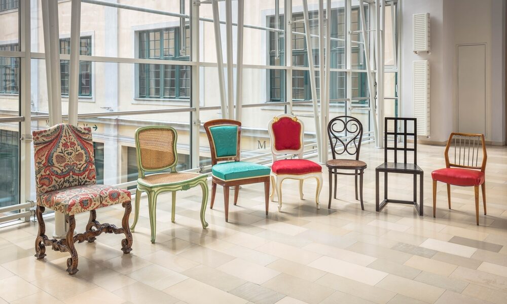 In einem hellen, modernen Raum mit großen Fenstern und gefliestem Boden sind sechs verschiedene Vintage-Stühle mit unterschiedlichen Designs und Bezügen aneinandergereiht.