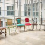 In einem hellen, modernen Raum mit großen Fenstern und gefliestem Boden sind sechs verschiedene Vintage-Stühle mit unterschiedlichen Designs und Bezügen aneinandergereiht.