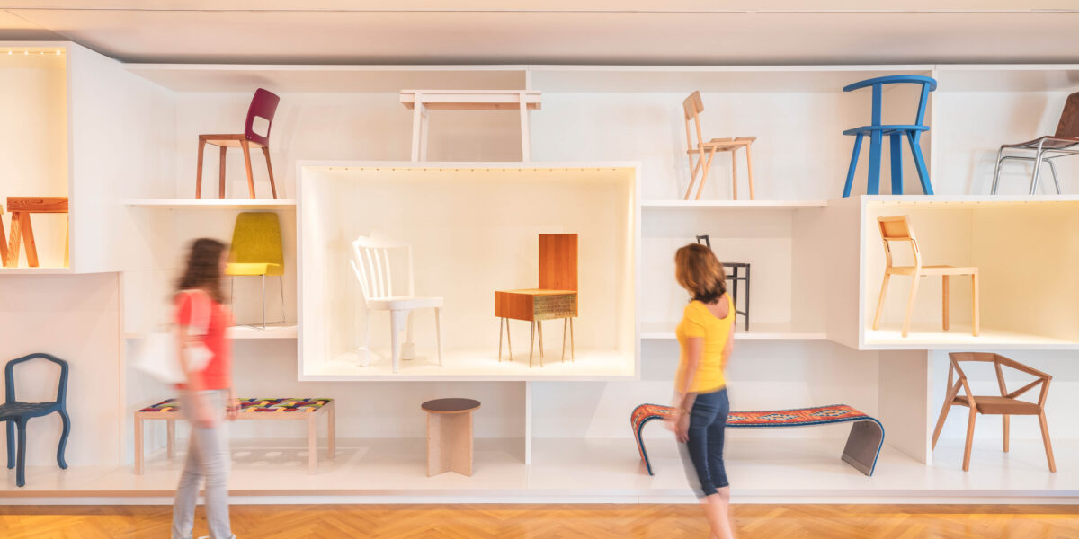 Zwei Personen gehen an einer Wand mit verschiedenen Stühlen und Bänken in einem hellen, modernen Galerieraum mit Holzfußboden vorbei.