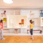 Zwei Personen gehen an einer Wand mit verschiedenen Stühlen und Bänken in einem hellen, modernen Galerieraum mit Holzfußboden vorbei.