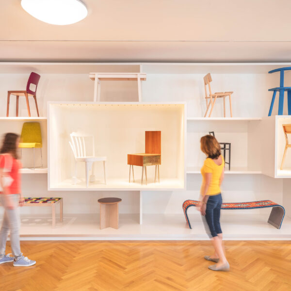 Zwei Personen gehen an einer Wand mit verschiedenen Stühlen und Bänken in einem hellen, modernen Galerieraum mit Holzfußboden vorbei.