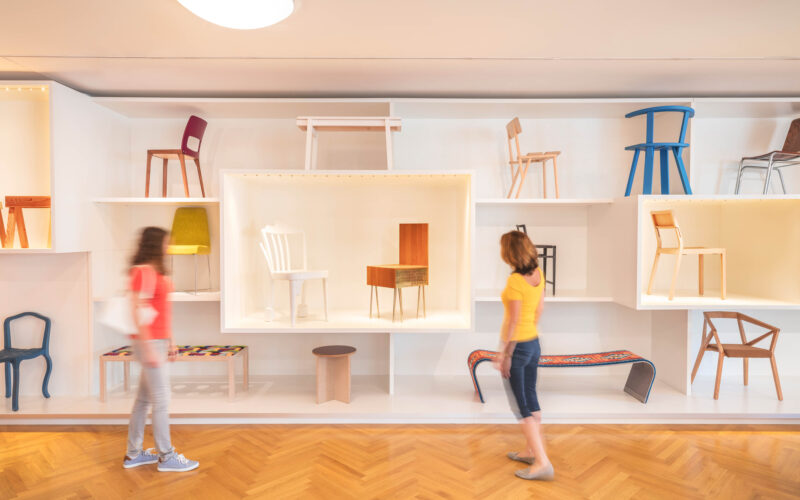 Zwei Personen gehen an einer Wand mit verschiedenen Stühlen und Bänken in einem hellen, modernen Galerieraum mit Holzfußboden vorbei.