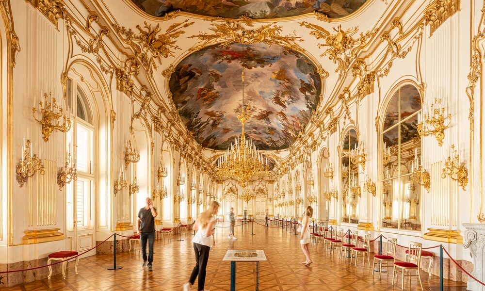 Verschnörkelter Ballsaal mit vergoldeten Dekorationen, großen Kronleuchtern, verspiegelten Wänden, bemalter Decke und mehreren Personen, die im großen Saal spazieren gehen oder Auslagen betrachten.