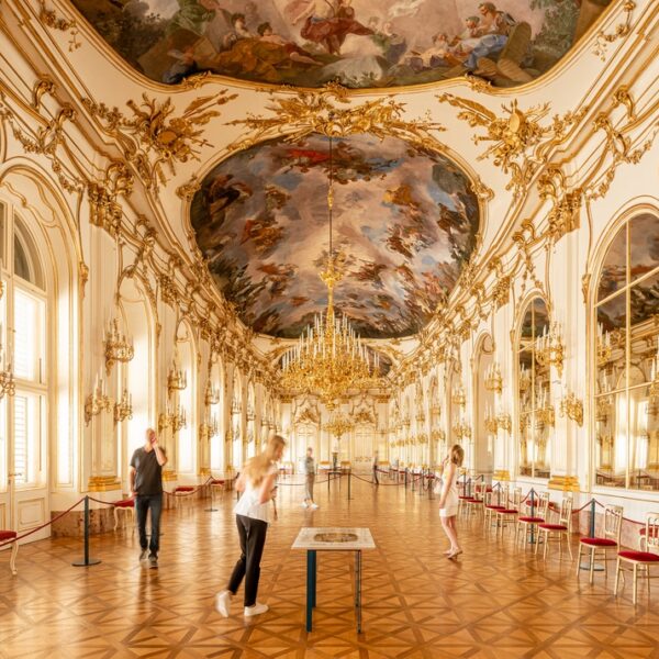 Verschnörkelter Ballsaal mit vergoldeten Dekorationen, großen Kronleuchtern, verspiegelten Wänden, bemalter Decke und mehreren Personen, die im großen Saal spazieren gehen oder Auslagen betrachten.