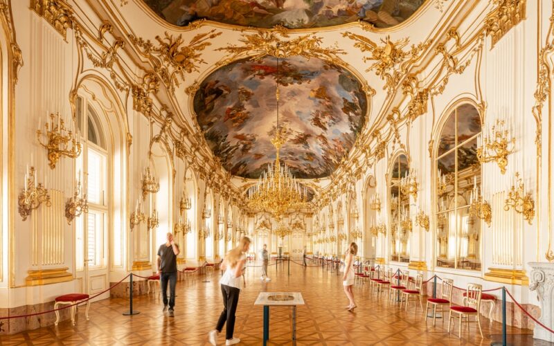 Verschnörkelter Ballsaal mit vergoldeten Dekorationen, großen Kronleuchtern, verspiegelten Wänden, bemalter Decke und mehreren Personen, die im großen Saal spazieren gehen oder Auslagen betrachten.