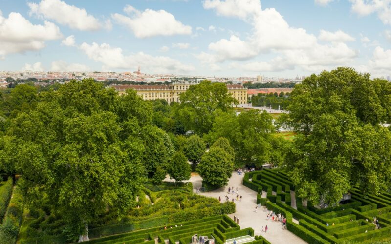Ein formaler Garten mit gepflegten Hecken, Kieswegen und großen Bäumen, mit einem historischen Palast und einer Stadtlandschaft im Hintergrund unter einem teilweise bewölkten Himmel.