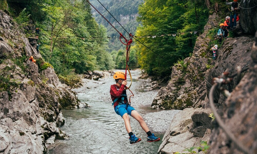 Eine Person, die einen Helm und ein Gurtzeug trägt, seilt sich über eine felsige Flussschlucht ab, umgeben von Bäumen und Bergen, während andere im Hintergrund warten.