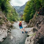 Eine Person, die einen Helm und ein Gurtzeug trägt, seilt sich über eine felsige Flussschlucht ab, umgeben von Bäumen und Bergen, während andere im Hintergrund warten.