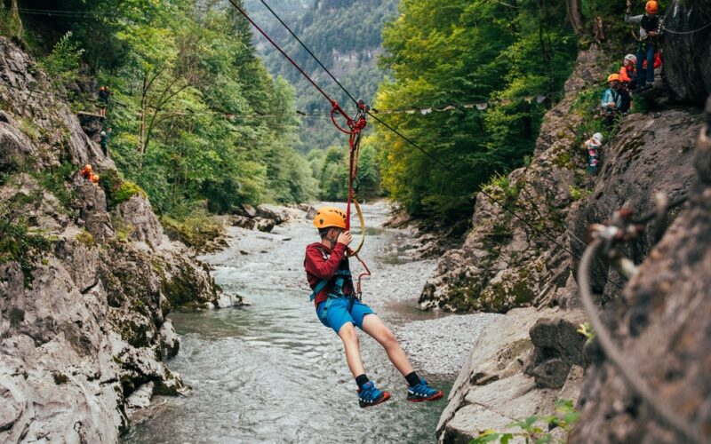 Eine Person, die einen Helm und ein Gurtzeug trägt, seilt sich über eine felsige Flussschlucht ab, umgeben von Bäumen und Bergen, während andere im Hintergrund warten.