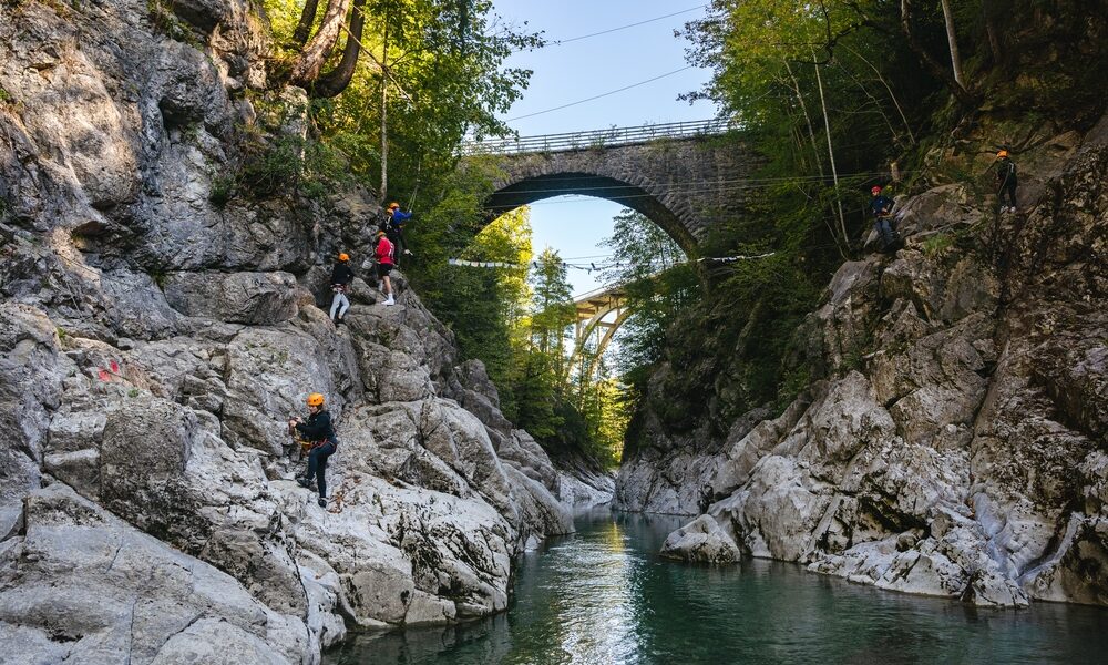 Menschen mit Helmen und Klettergurten klettern an felsigen Canyonwänden über einem Fluss, über denen zwei Brücken inmitten von Bäumen und Sonnenlicht zu sehen sind.