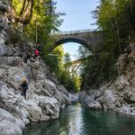 Menschen mit Helmen und Klettergurten klettern an felsigen Canyonwänden über einem Fluss, über denen zwei Brücken inmitten von Bäumen und Sonnenlicht zu sehen sind.