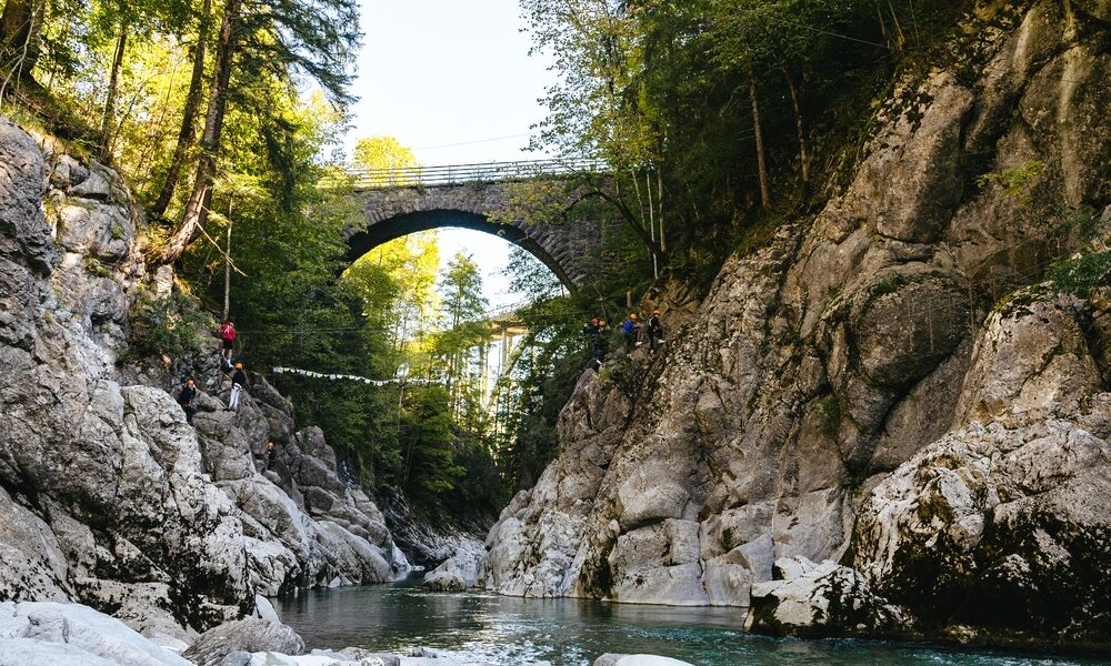 Person beim Ziplining über eine felsige Flussschlucht mit einer Steinbogenbrücke und hohen Bäumen im Hintergrund an einem sonnigen Tag.