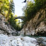Person beim Ziplining über eine felsige Flussschlucht mit einer Steinbogenbrücke und hohen Bäumen im Hintergrund an einem sonnigen Tag.