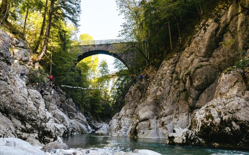 Person beim Ziplining über eine felsige Flussschlucht mit einer Steinbogenbrücke und hohen Bäumen im Hintergrund an einem sonnigen Tag.
