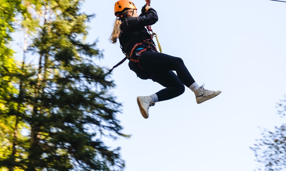 Eine Person, die einen Helm und ein Gurtzeug trägt, fährt auf einer Seilrutsche durch ein bewaldetes Gebiet unter freiem Himmel.