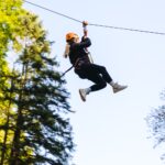 Eine Person, die einen Helm und ein Gurtzeug trägt, fährt auf einer Seilrutsche durch ein bewaldetes Gebiet unter freiem Himmel.