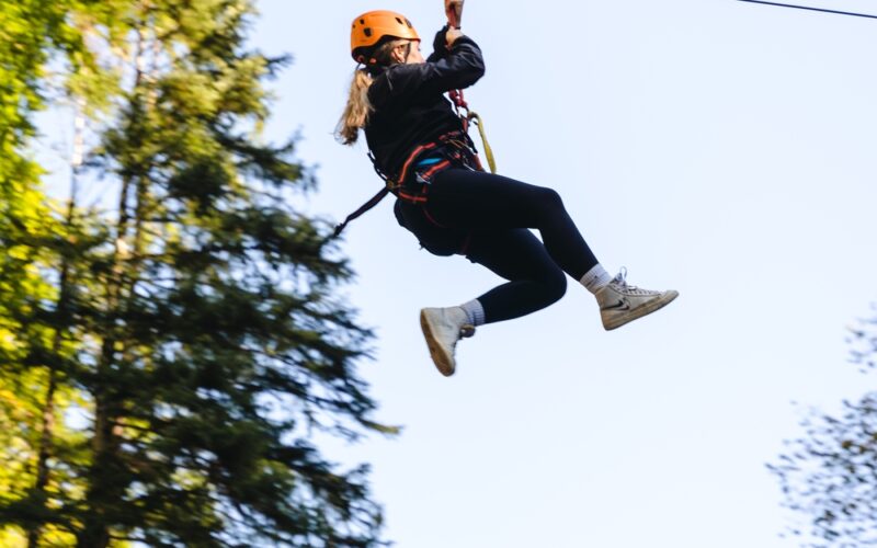 Eine Person, die einen Helm und ein Gurtzeug trägt, fährt auf einer Seilrutsche durch ein bewaldetes Gebiet unter freiem Himmel.