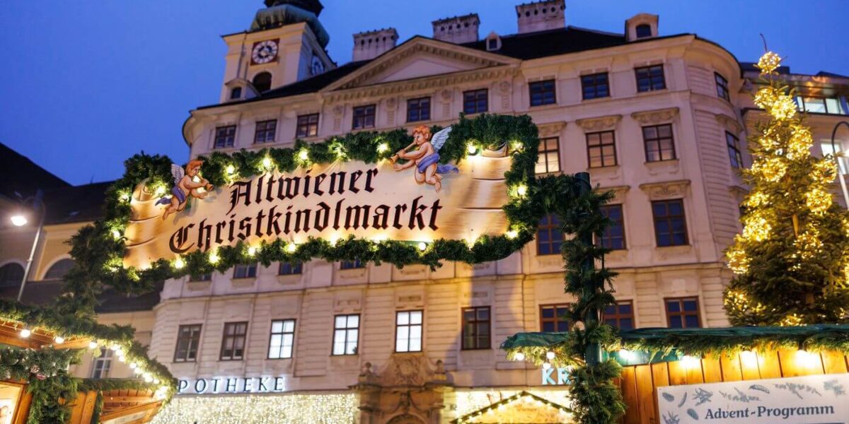 Ein beleuchtetes Schild mit der Aufschrift "Altwiener Christkindlmarkt" auf einem traditionellen Weihnachtsmarkt, mit geschmückten Ständen und einem historischen Gebäude im Hintergrund.
