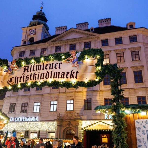 Ein beleuchtetes Schild mit der Aufschrift "Altwiener Christkindlmarkt" auf einem traditionellen Weihnachtsmarkt, mit geschmückten Ständen und einem historischen Gebäude im Hintergrund.