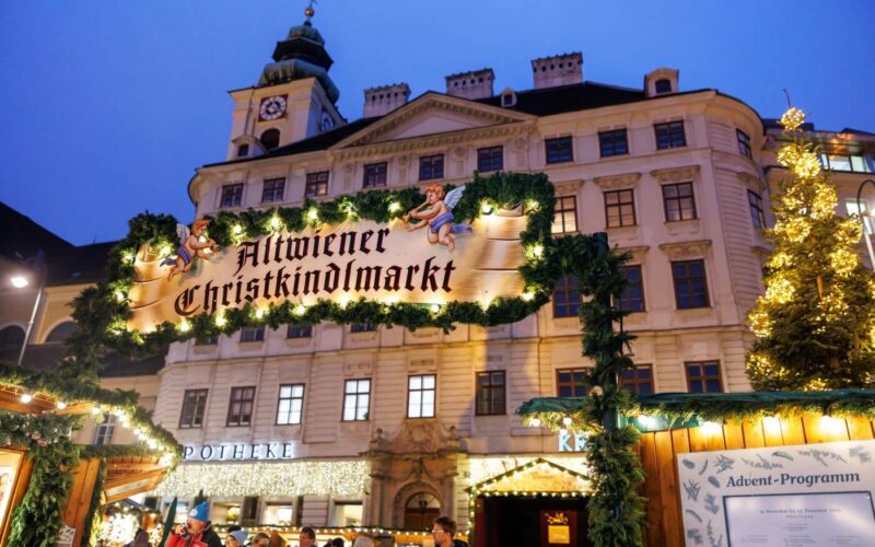 Ein beleuchtetes Schild mit der Aufschrift "Altwiener Christkindlmarkt" auf einem traditionellen Weihnachtsmarkt, mit geschmückten Ständen und einem historischen Gebäude im Hintergrund.