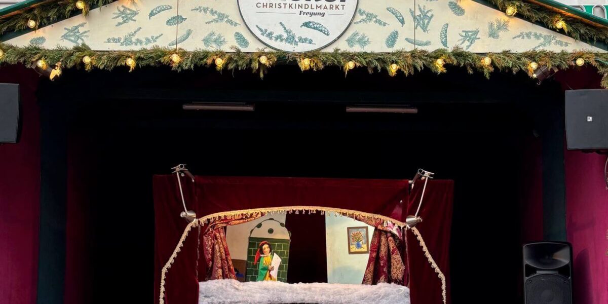 Auf der Freilichtbühne des Alt Wiener Christkindlmarkts mit geschmückter Fassade und festlicher Beleuchtung über der Bühne sehen Kinder ein Puppentheater.