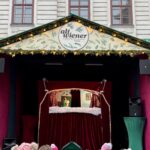 Auf der Freilichtbühne des Alt Wiener Christkindlmarkts mit geschmückter Fassade und festlicher Beleuchtung über der Bühne sehen Kinder ein Puppentheater.