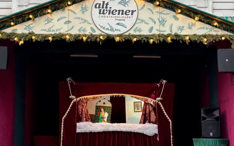 Auf der Freilichtbühne des Alt Wiener Christkindlmarkts mit geschmückter Fassade und festlicher Beleuchtung über der Bühne sehen Kinder ein Puppentheater.