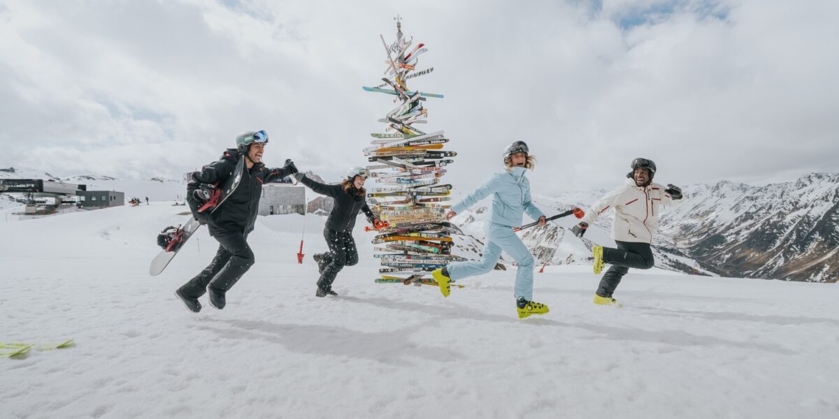 Vier Personen in Skikleidung halten sich an den Händen und laufen im Kreis um einen Weihnachtsbaum aus Skiern auf einem verschneiten Berg mit bewölktem Himmel und fernen Gipfeln.