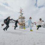 Vier Personen in Skikleidung halten sich an den Händen und laufen im Kreis um einen Weihnachtsbaum aus Skiern auf einem verschneiten Berg mit bewölktem Himmel und fernen Gipfeln.
