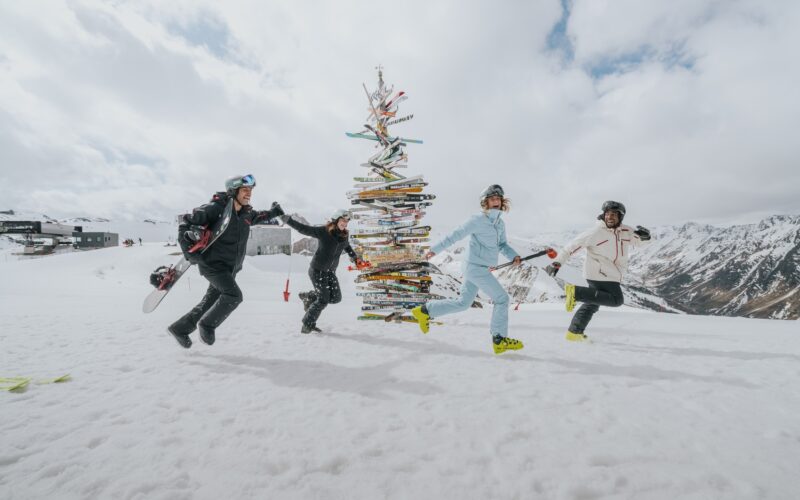 Vier Personen in Skikleidung halten sich an den Händen und laufen im Kreis um einen Weihnachtsbaum aus Skiern auf einem verschneiten Berg mit bewölktem Himmel und fernen Gipfeln.