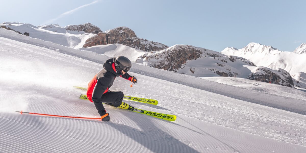 Person beim Skifahren auf einer präparierten Schneepiste, die sich in eine scharfe Kurve neigt, während der Schnee aufspritzt, im Hintergrund sind die Berge zu sehen.