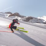 Person beim Skifahren auf einer präparierten Schneepiste, die sich in eine scharfe Kurve neigt, während der Schnee aufspritzt, im Hintergrund sind die Berge zu sehen.