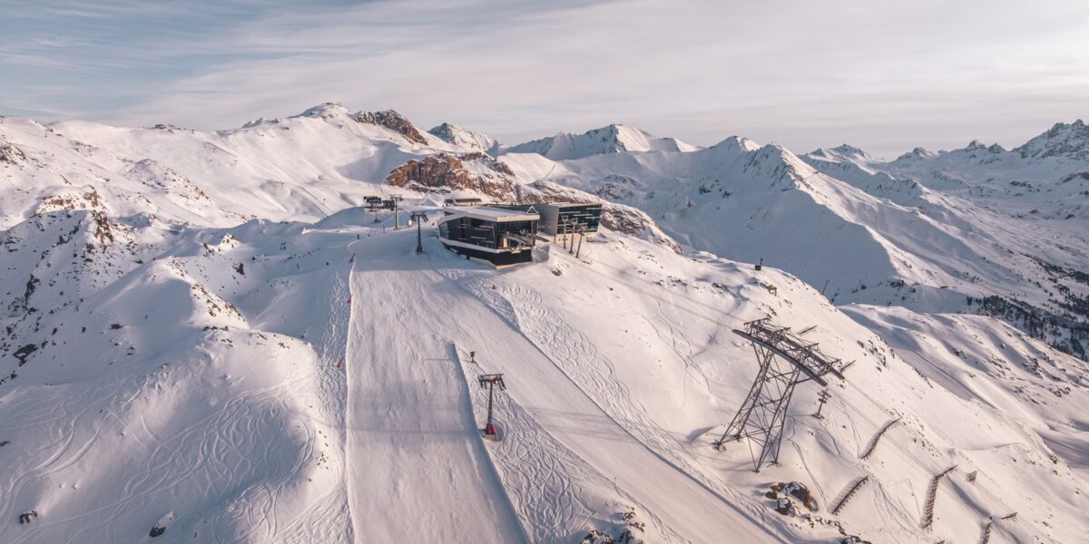 Luftaufnahme eines schneebedeckten Berghangs mit einer Skiliftstation, Skispuren und schroffen Gipfeln im Hintergrund unter einem bewölkten Himmel.