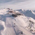 Luftaufnahme eines schneebedeckten Berghangs mit einer Skiliftstation, Skispuren und schroffen Gipfeln im Hintergrund unter einem bewölkten Himmel.
