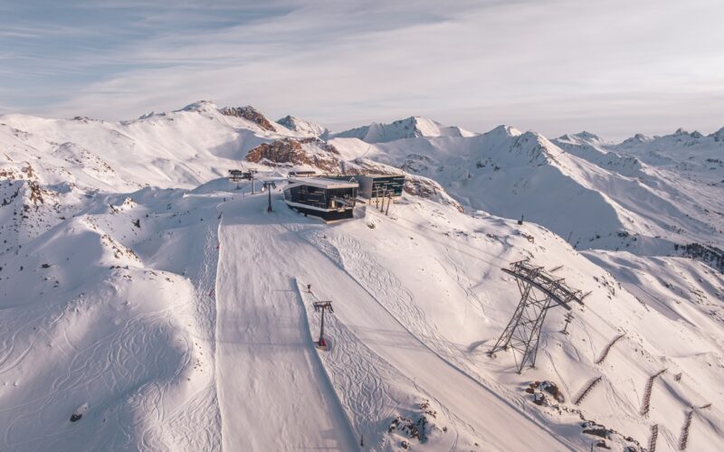 Luftaufnahme eines schneebedeckten Berghangs mit einer Skiliftstation, Skispuren und schroffen Gipfeln im Hintergrund unter einem bewölkten Himmel.