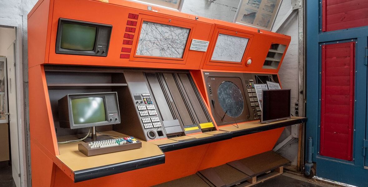 Große orangefarbene Steuerkonsole im Vintage-Stil mit mehreren Bildschirmen, Tastaturen, Tasten und Karten vor einer weißen Wand in einem Museum oder einer industriellen Umgebung.
