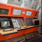 Große orangefarbene Steuerkonsole im Vintage-Stil mit mehreren Bildschirmen, Tastaturen, Tasten und Karten vor einer weißen Wand in einem Museum oder einer industriellen Umgebung.