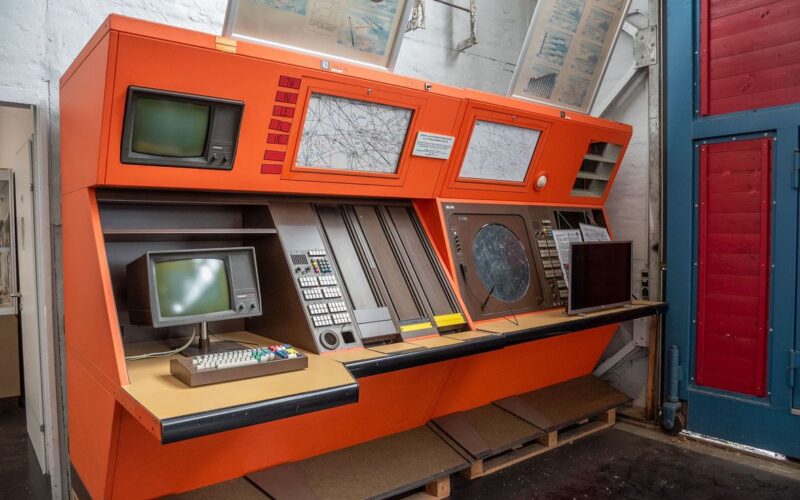 Große orangefarbene Steuerkonsole im Vintage-Stil mit mehreren Bildschirmen, Tastaturen, Tasten und Karten vor einer weißen Wand in einem Museum oder einer industriellen Umgebung.
