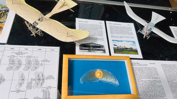Vitrine mit Modellen früher Flugmaschinen, Informationsblättern, einem gerahmten flügelförmigen Objekt und einem Zeitdiagramm, die alle mit der Geschichte der Luftfahrt zu tun haben.