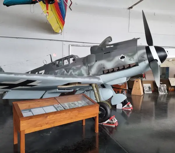 Ein deutsches Kampfflugzeug aus dem Zweiten Weltkrieg ist in einem Museumshangar ausgestellt, in dessen Nähe sich Informationstafeln und Artefakte befinden.