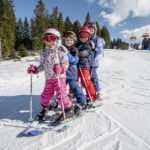 Vier Kinder in Skiausrüstung stehen auf Skiern in einer Reihe auf einer verschneiten Piste in der Nähe eines Skilifts, mit Bäumen und einem blauen Himmel im Hintergrund.