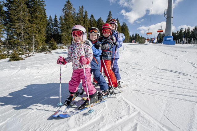 Vier Kinder in Skiausrüstung stehen auf Skiern in einer Reihe auf einer verschneiten Piste in der Nähe eines Skilifts, mit Bäumen und einem blauen Himmel im Hintergrund.