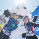 Zwei Personen in Wintersportkleidung posieren mit einem großen weißen Hundemaskottchen im Freien im Schnee, während die Sonne hell über ihnen scheint.