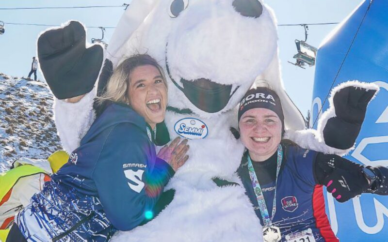Zwei Personen in Wintersportkleidung posieren mit einem großen weißen Hundemaskottchen im Freien im Schnee, während die Sonne hell über ihnen scheint.