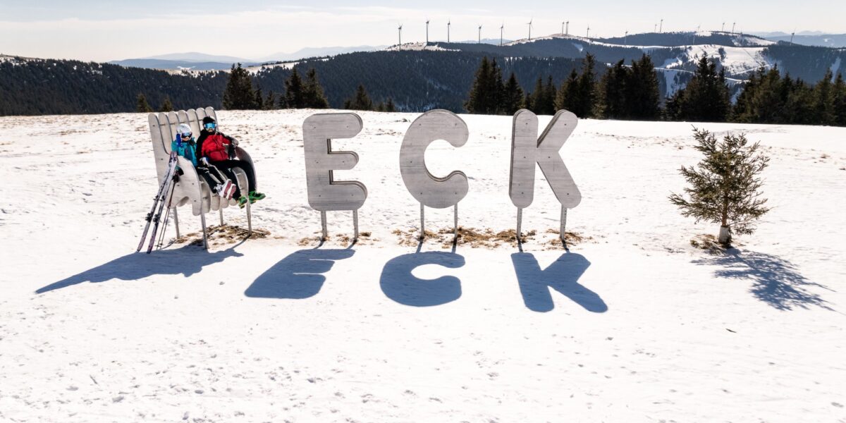 Eine Person in Skikleidung sitzt auf einer Bank neben großen Metallbuchstaben mit der Aufschrift "ECK" auf einem verschneiten Hügel, im Hintergrund sind Bäume und Windräder zu sehen.