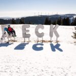 Eine Person in Skikleidung sitzt auf einer Bank neben großen Metallbuchstaben mit der Aufschrift "ECK" auf einem verschneiten Hügel, im Hintergrund sind Bäume und Windräder zu sehen.