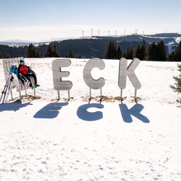 Eine Person in Skikleidung sitzt auf einer Bank neben großen Metallbuchstaben mit der Aufschrift "ECK" auf einem verschneiten Hügel, im Hintergrund sind Bäume und Windräder zu sehen.