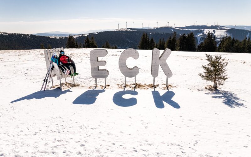 Eine Person in Skikleidung sitzt auf einer Bank neben großen Metallbuchstaben mit der Aufschrift "ECK" auf einem verschneiten Hügel, im Hintergrund sind Bäume und Windräder zu sehen.